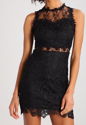 Vestido mini negro sin mangas de encaje con estampados florales, escote transparente y corte en la cintura, llevado por una persona de pie.