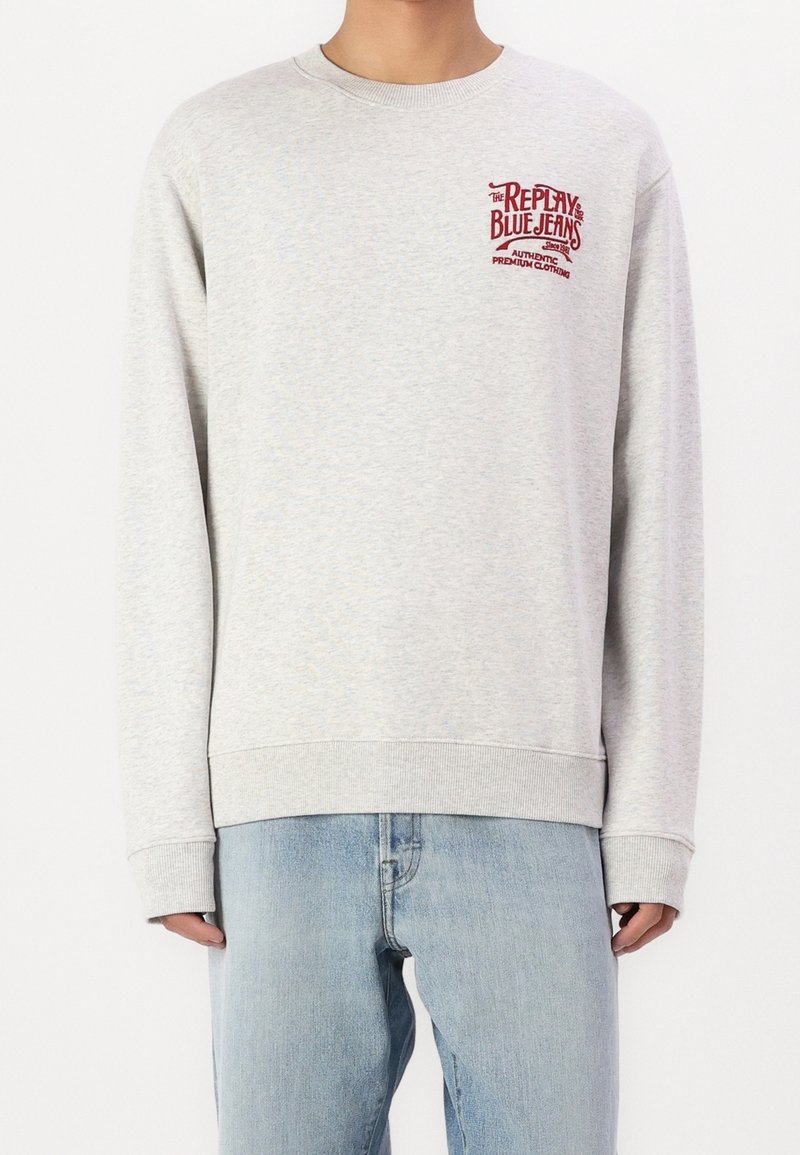 Sweat-shirt gris clair à manches longues avec logo rouge "Replay Blue Jeans" sur la poitrine, porté avec un jean bleu clair.