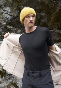 Svart kortärmad skjorta i kombination med beige ytterplagg och mörka byxor. Modellen bär en gul stickad mössa, vilket visar en avslappnad utomhusstil.