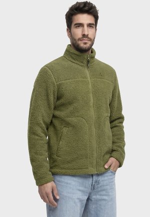Mann trägt grüne Fleecejacke mit Reißverschluss und hohem Kragen sowie hellblaue Jeans und steht vor schlichtem Hintergrund.