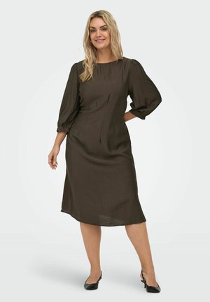 Smilende kvinne iført en mørk brun midi-kjole med puffede trekvart ermer og svarte slingback-hæler, stående mot en hvit bakgrunn.