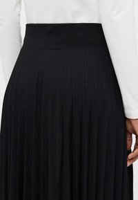 Svart plisserad kjol med ett slätt, brett midjeband. Tyget har en mjuk textur och når till mitten av vadarna.