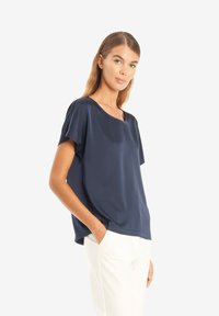 Mörkblå blus med kort ärm i slät tyg, med vid halsringning, avslappnad passform och sprund i sidorna för ökad rörlighet.