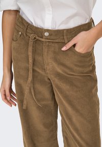 Gros plan d'une personne portant un pantalon en velours côtelé marron avec une ceinture en tissu, une main dans la poche, associé à une chemise blanche.