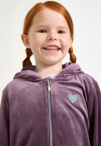 Ragazza sorridente dai capelli rossi con lentiggini, che indossa una felpa viola con zip e un piccolo cuore ricamato sul petto, su uno sfondo neutro.