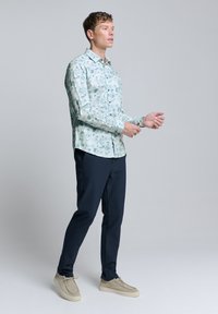 Lange mouw shirt in lichtblauw met een bloemenpatroon, gedragen met donkere broek en beige schoenen. Heeft een knoopsluiting en een kraag.