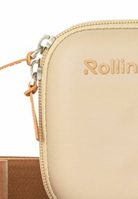 Gros plan sur une pochette beige zippée avec des tirettes de fermeture éclair couleur tan et une partie de la marque « Rollin » sur une surface lisse.