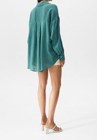 Woman wearing a loose teal long-sleeve shirt, white shorts, and white heeled sandals, standing against a plain light background.