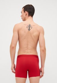 Röda bomullsboxershorts med en åtsittande design; modellens rygg har en dekorativ svart tatuering av en lilja.