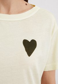 Lichtgele katoenen t-shirt met een donkergroen hartontwerp op de linkerborst. Ribgebreide halslijn en korte mouwen. Zachte textuur.