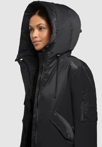 Black padded jacket with a large hood and zip front. Features a chest pocket and a side pocket with a flap. Smooth, shiny fabric texture.