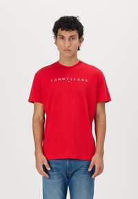 T-shirt rouge en coton avec encolure ras du cou. Affiche le texte blanc "TOMMYJEANS" sur la poitrine. Porté avec un jean bleu. Design simple à manches courtes.