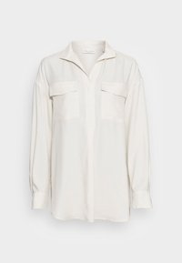Blusa blanca de manga larga con cuello, dos bolsillos en el pecho y un corte relajado. Tela suave con un ligero brillo, dobladillo curvado.