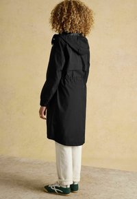 Čierny nepremokavý kabát s kapucňou, s uťahovacím pásom a dlhými rukávmi, v kombinácii s bielymi nohavicami a zelenými teniskami. Textúrované pozadie.