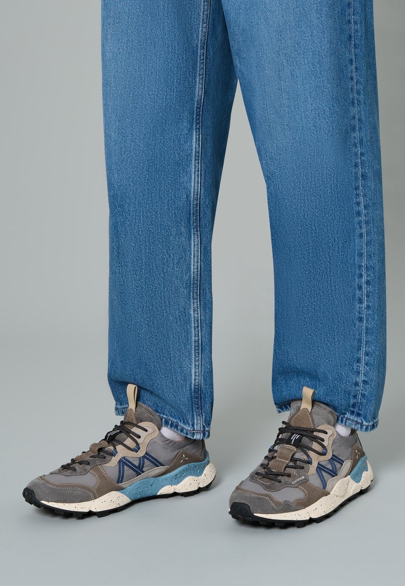 Baskets grises et bleues avec un dessus texturé ; présentent des accents en maille et en caoutchouc, des lacets noirs et une semelle mouchetée. Portées avec un jean bleu.