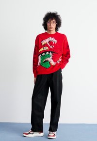 Un suéter rojo con un diseño de monstruo colorido, combinado con pantalones negros holgados y zapatillas rojas y blancas. El fondo es liso y de color claro.
