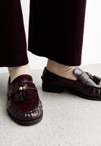 Bordeaux leren instappers met een decoratieve kwast, met een ronde neus en textuursteek, gecombineerd met beige sokken en brede corduroy broeken.