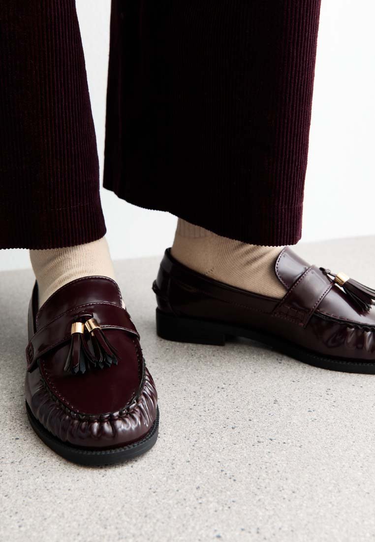 Bordeaux leren instappers met een decoratieve kwast, met een ronde neus en textuursteek, gecombineerd met beige sokken en brede corduroy broeken.