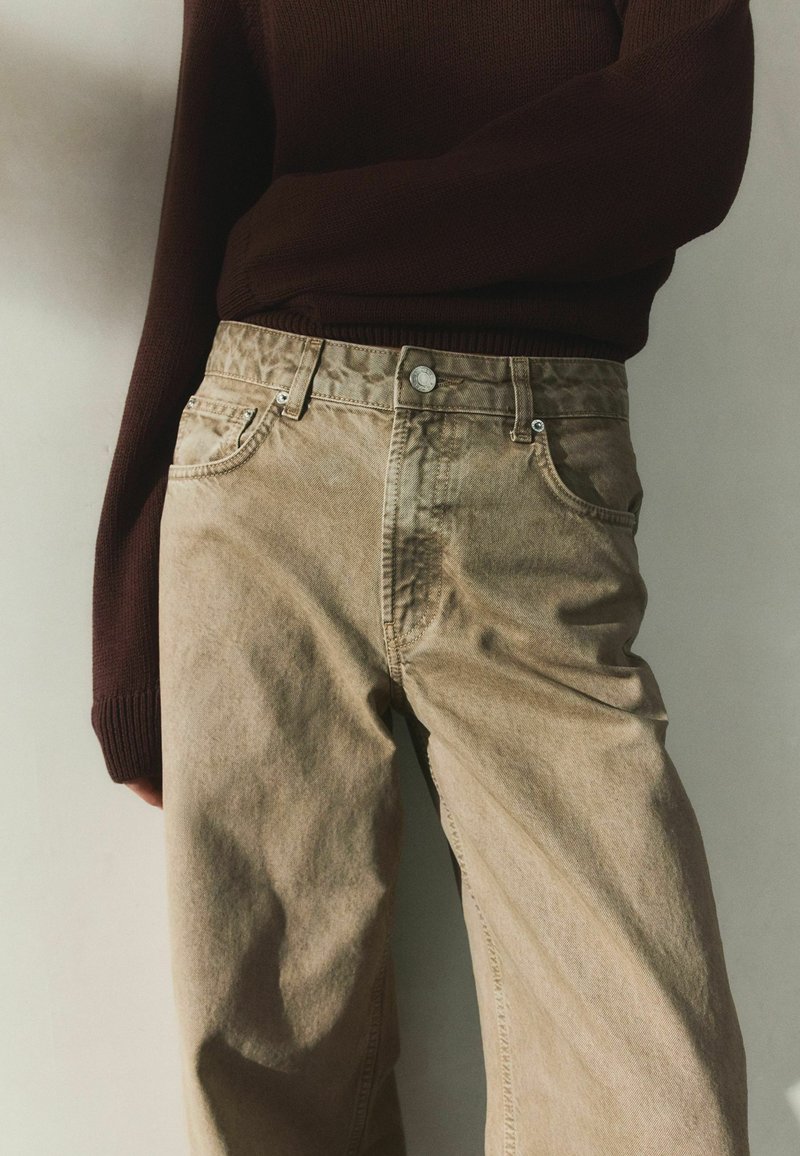 Person iført beige bukser med høj talje og en mørkebrun langærmet sweater, stående foran en ensfarvet lys baggrund.