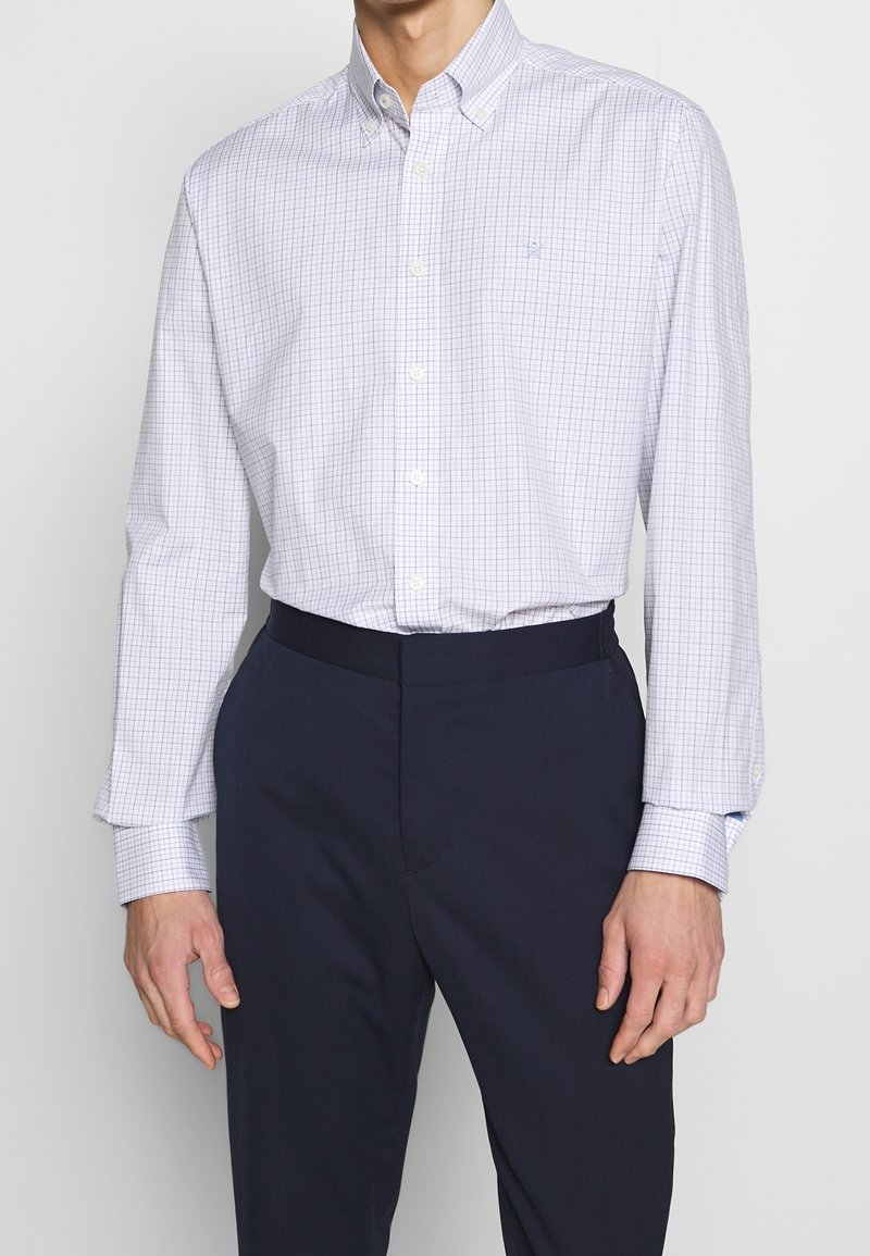 Homme portant une chemise blanche à manches longues avec un motif à carreaux bleus et un pantalon bleu marine foncé, debout devant un fond uni.