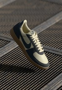 Sneaker Nike con una toma superiore crema, pannelli con accenti blu navy e suola in gomma. Design testurizzato con lacci piatti e logo sulla linguetta.