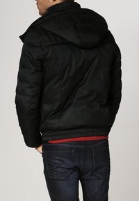 Chaqueta acolchada negra con capucha, con dobladillo ajustado y textura suave. Lleva puesta una camisa roja y la combina con vaqueros oscuros.