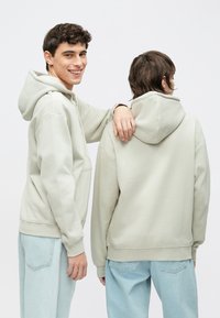 Šviesiai žali gobtuvai, pagaminti iš minkštos medžiagos, su kengūros kišene, gofruotomis rankogaliais ir matomomis siūlėmis, dėvimi su šviesiai mėlynomis kelnėmis.