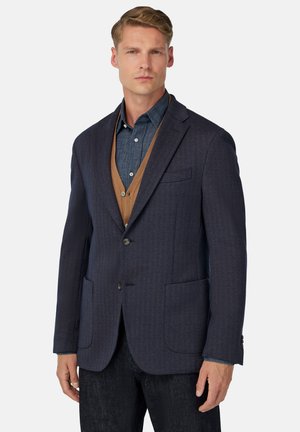Marineblauer Fischgrät-Blazer mit zwei Fronttaschen, Ein-Knopf-Verschluss und darüber trägt er eine braune Weste über einem blauen Hemd.