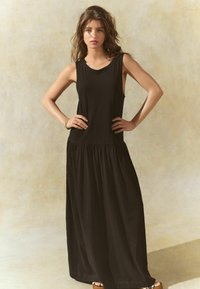 Robe maxi noire en tissu doux, avec un design sans manches, une taille froncée, une jupe fluide et une coupe au niveau des chevilles. Texture lisse.