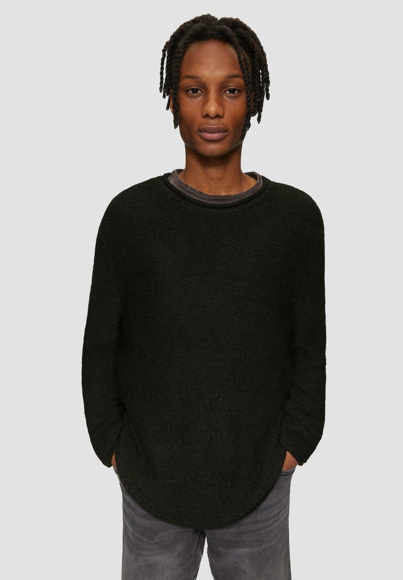 Schwarzer Strickpullover mit rundem Ausschnitt, lockerem Schnitt, strukturierter Jerseystoff und leicht verlängertem Rückensaum. Getragen über einem grauen Hemd.