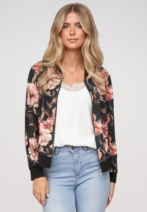 Joven con cabello rubio largo, vestida con una chaqueta bomber negra con flores sobre una camiseta blanca con encaje y unos vaqueros azul claro, de pie frente a un fondo liso.