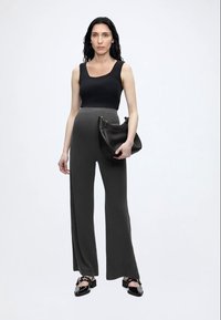 Γυναίκα με μακριά μαύρα μαλλιά φοράει μαύρο αμάνικο τοπ, ψηλόμεσο σκούρο γκρι παντελόνι με φαρδιά πατζάκια, μαύρα μυτερά παπούτσια και κρατάει μαύρη τσάντα.