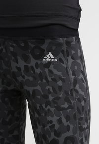Leggings neri con un motivo a stampa leopardata grigia, caratterizzati da una texture liscia e cuciture a zig-zag. Il logo Adidas è visibile sulla coscia.