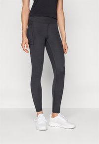 Mujer vestida con leggings negros con un bolsillo lateral y zapatillas deportivas blancas, de pie contra un fondo claro y liso.