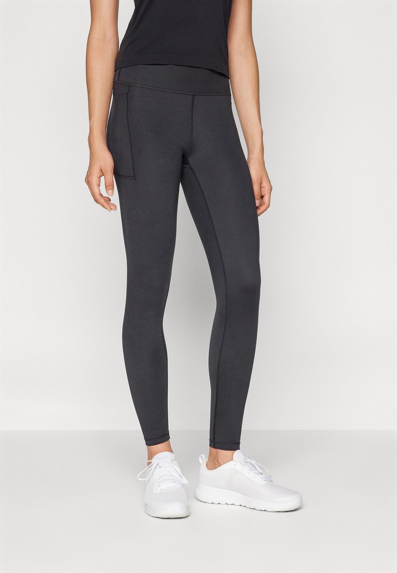 Mujer vestida con leggings negros con un bolsillo lateral y zapatillas deportivas blancas, de pie contra un fondo claro y liso.