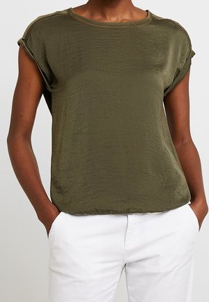 Oliwkowy top z krótkim rękawem o teksturowanym wykończeniu, z podwiniętymi rękawami i okrągłym dekoltem. Połączony z białymi spodniami dla kontrastu.