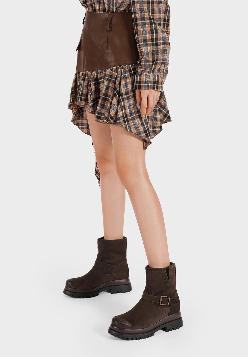 Stivali ankle in suede marrone con punta arrotondata, suola piatta e dettagli decorativi con fibbia. Tessuto texture e cuciture visibili.