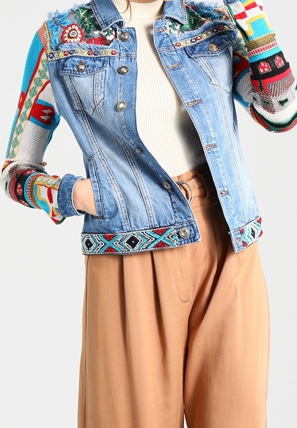 Farkkutakki, jossa on värikkäitä brodeerattuja kukka- ja geometrisia kuvioita hihoissa ja helmassa, puettuna valkoisen ribbipaidan ja vaaleanruskeiden leveälahkeisten housujen päällä.