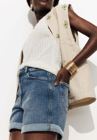 Shorts en denim avec une teinte bleu délavé, ourlet retroussé, associés à un haut en tricot blanc texturé et un gilet sans manches beige décoré d'accents verts.