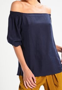 Navy off-the-shoulder blouse with elastic smocking at the neckline, short puff sleeves, and a relaxed fit. Smooth, lightweight fabric.