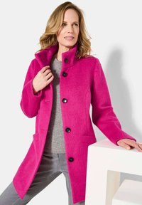 Fuchsia Wolle-Mantel mit hohem Kragen, fünf schwarzen Knöpfen und seitlichen Taschen. Kombiniert mit einem grauen Pullover und vor einem weißen Hintergrund gezeigt.