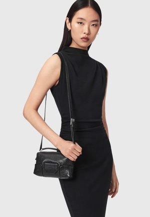 Femme portant une robe noire sans manches tenant un petit sac en cuir noir texturé porté en bandoulière avec une longue sangle.