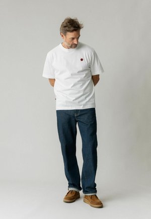 Mand med kort brunt hår og skæg iført hvid t-shirt, mørkeblå jeans og brune sko, står med hænderne bag ryggen.