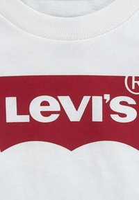 Bela bombažna majica s kratkimi rokavi z velikim rdečim logotipom Levi's. Dizajn vključuje značilno ukrivljeno obliko in debele bele črke na rdeči podlagi.