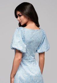 Robe en dentelle florale bleu clair avec des manches bouffantes, silhouette ajustée et décolleté carré, présentant un motif complexe et une finition texturée.