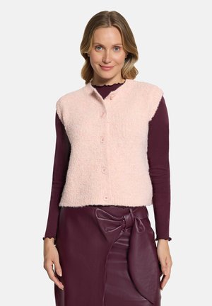 Woman wearing a light pink buttoned sleeveless vest over a long-sleeve burgundy top and a burgundy leather skirt with a large bow detail.