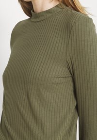 Olivegrünes, geripptes Longsleeve-Oberteil mit Stehkragen. Der Stoff hat eine glatte Textur und vertikale Streifen, die entlang der Oberfläche verlaufen.