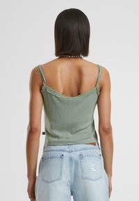 Groene geribbelde tanktop met kantafwerking, met een lage rugontwerp. G gedragen met lichtblauwe high-waisted jeans, wat de casual stijl benadrukt.