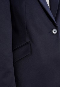 Námořnický blazer s diagonální texturou, zapínáním na jeden knoflík a náprsní kapsou. Látka vypadá hladce a strukturovaně.