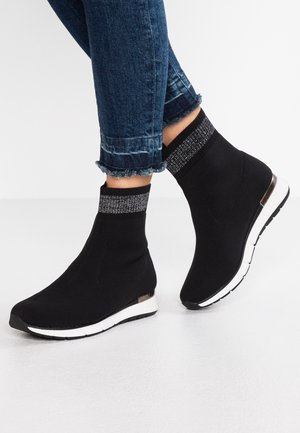 Sorte ankelsneakers i sokkestil med hvide såler og sølvskimrende detaljer ved kanterne, båret med blå jeans med frynset kant.